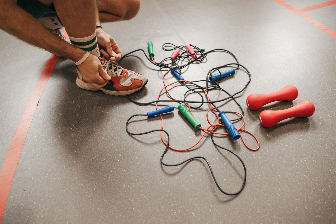 services-02 Individual tying shoelaces near colorful jump ropes and red dumbbells on a gym floor.