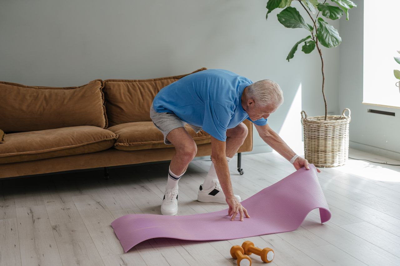 services-03 An elderly man sets up his yoga mat indoors, embodying a healthy lifestyle.